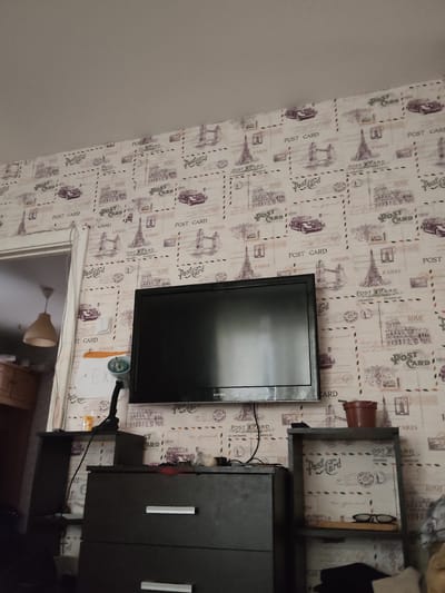 The image shows a room with a television mounted on a wall covered in wallpaper featuring vintage postcard designs. To the left of the television, a doorway reveals a glimpse of another room with a pendant light fixture and furniture. Below the television, a dark dresser with silver drawer pulls is visible. To the right of the television, a small shelf holds a brown pot and a pair of glasses. The scene depicts a domestic interior, possibly a living room or bedroom. There are no people or animals visible in the image. The location is specified as Андрэеўшчынскі сельскі Савет, Belarus.