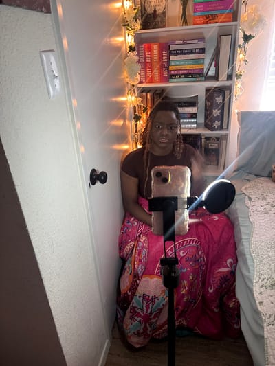 The image shows a woman sitting on the floor in front of a bookshelf. She is positioned to the side, facing forward, holding a cell phone with a ring light attached to it, which is positioned in front of her. The bookshelf is full of books and is decorated with artificial flowers and string lights. There is a bed in the background. The room appears to be a bedroom. There is a door knob and light switch in the foreground. The exact location within Arlington, United States of America, cannot be determined from the image.