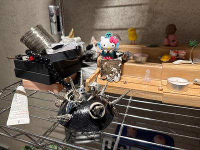An extreme close-up, eye-level shot shows a collection of various objects on and around a multi-tiered metal shelving unit. The objects include a small black sphere adorned with silver spikes and rings, a tube of white cream with text, a stack of black boxes topped with a silver bracelet and a strand of black beads with red and white accents, a Hello Kitty figurine wearing a crown and dress, a yellow chick figurine, a small pink figurine of a seated person, a green frog figurine, two small pink Hello Kitty figurines, a yellow hat, a small white ceramic object, a lit tealight candle in a silver holder, and other small decorative items. The background consists of a textured wall and wooden shelves. The scene appears to be an indoor setting, possibly a home or a shop. The shelving unit is made of metal rods and is partially filled with these various items.