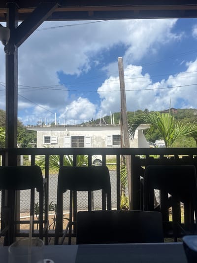 This image depicts a view from a covered porch or patio located in Olde Yard Village, British Virgin Islands. The foreground features a partial view of a dark table surface on the bottom with a clear glass and straw visible on the left. Three dark, high-backed chairs or bar stools are silhouetted in the mid-foreground, positioned behind a dark wooden railing composed of horizontal top and bottom rails connected by vertical slats.
Beyond the railing, a chain-link fence runs horizontally above an asphalt road. To the left of the road, a white car is partially visible, parked on a grassy verge. Central to the mid-ground is a light-colored, possibly stucco or concrete, single-story building featuring multiple rectangular windows equipped with louvered shutters, some appearing open, others closed. The building's roof line shows several vertical white pipes or vents. Green tropical vegetation, including palm fronds, is present on both sides of the road and around the building. A tall, weathered wooden utility pole stands prominently on the right side, extending from the mid-ground into the sky, with multiple black power lines traversing the upper frame.
The background consists of lush green, vegetation-covered hills rising under a blue sky dotted with scattered white cumulus clouds. Distant structures with reddish-brown roofs are visible on the hillsides. No people, specific actions, or interactions are observable. The scene is illuminated by daylight.