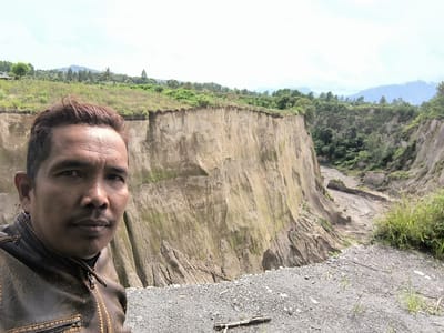 This is a medium shot, outdoor, selfie-style photograph featuring a man in the foreground. The man, who appears to be of Southeast Asian descent, has short, dark hair and a mustache. He is wearing a dark brown leather jacket with a zipper and intricate stitching. He is looking directly at the camera with a neutral expression.

Behind the man is a dramatic landscape characterized by steep, eroded cliffs and a deep ravine. The cliffs are a light tan color, with streaks of green vegetation clinging to their surfaces. The ravine floor appears to be a mix of sand, gravel, and dried mud, with evidence of water flow. The background features rolling hills covered in lush green vegetation and trees, with mountains visible in the distance under an overcast sky.

The overall impression is one of a rugged and natural environment. The lighting suggests it is daytime, likely overcast, as there are no strong shadows. The location appears to be rural or natural, possibly in a mountainous or volcanic region given the geological formations. The image was taken in Kebunbaru, Indonesia. No text is visible in the image.