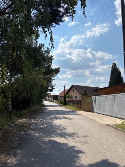 The image depicts a residential street in Červená Píska, Czechia, on a sunny day with scattered clouds. On the left side of the asphalt road, there is a dense line of trees and vegetation. The road itself is mostly in shadow due to the trees. On the right side of the road, there are houses with tiled roofs and fences. A large grey metal gate is visible in the foreground on the right. Power lines run diagonally across the blue sky. The overall impression is a quiet, suburban street scene.