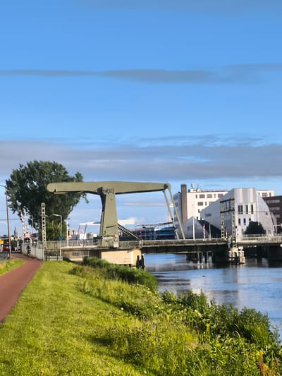The image depicts a drawbridge over a canal in Alkmaar, Netherlands. The drawbridge, painted a muted green, is positioned horizontally across the waterway. On the left side of the canal, a paved red path runs parallel to the grassy bank. A person on a bicycle is visible on this path, heading away from the viewer. In the background, several buildings are present, including a white, multi-story structure with red-paned windows. The sky is blue with wispy clouds. The overall scene is one of urban infrastructure set against a natural environment.