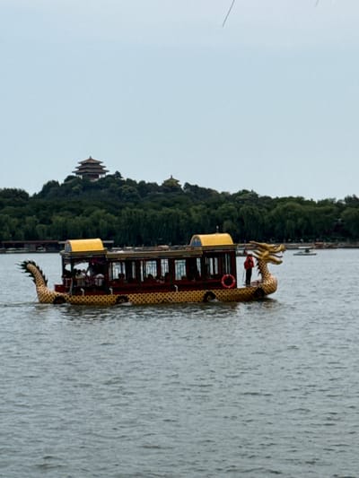 A dragon boat is sailing on a lake in front of a hill covered with trees, with a pagoda on top. The boat has a dragon head at the front and a dragon tail at the back, both made of gold. The boat is decorated with red and yellow colors. There are people inside the boat. The sky is overcast, suggesting it might be a cloudy day. The image is taken from a landscape perspective. The location is Xicheng District, China.