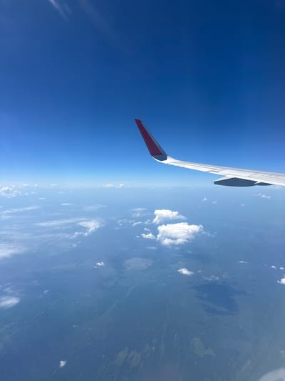 The image captures a view from an airplane window, looking down at the Earth. The most prominent subject is the wing of the airplane, which extends from the left side of the frame into the upper portion of the image. The wing is white with a red and white tip and appears to be in good condition. Below the wing, the landscape of Russia unfolds. The terrain is characterized by a vast expanse of land, possibly forest or tundra, punctuated by numerous lakes and rivers, reflecting the name "Многоозёрный" (meaning "Many-lakes"). Patches of white clouds are scattered across the sky and hover over the land, suggesting fair weather. The sky is a deep, clear blue, indicating it is daytime, likely late morning or early afternoon due to the bright illumination. The horizon line is visible, marking the boundary between the sky and the Earth. There is no visible text in the image. The scene evokes a sense of travel, journey, and the vastness of the natural world.