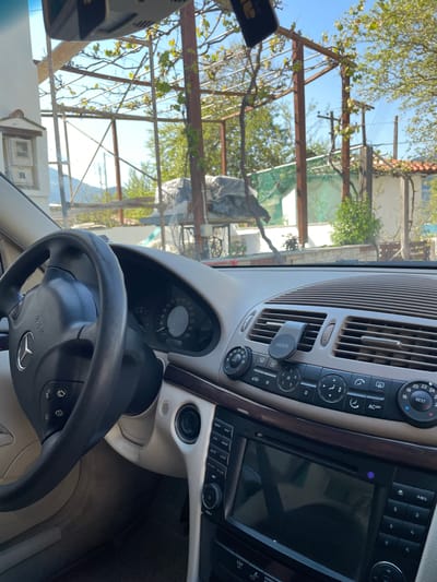 The image captures the interior of a car, specifically the dashboard and steering wheel, looking out through the windshield towards an outdoor scene. The car appears to be a Mercedes-Benz, evidenced by the logo on the steering wheel and the design of the console. The steering wheel has "SRS AIRBAG" printed on it. The dashboard features a speedometer with markings up to 260 km/h, and an infotainment system with buttons labeled "MENU," "NAVI," "DVD," "MODE," "MUTE," "LOUD," "GPS CARD," "SD CARD," "RST," and "VOL+." Below these are numeric buttons "1" through "6" and "AMS." Climate control dials show temperatures "16," "18," "20," "22," "24," "REST," and "AUTO," along with an "AC OFF" button. An "ESP OFF" button is also visible. A "VIDVIE" brand phone holder is clipped to an air vent.

The activity depicted is a static view from a parked car. Outside, under a clear blue sky, green foliage and tree branches are visible, suggesting lush vegetation. A large metal pergola or structural framework, possibly for vines, is prominent, with grapevines (or similar) growing on it. In the background, power lines or utility pylons can be discerned, along with parts of a building or house and some objects covered in tarps. The bright sunlight indicates it is daytime, likely a sunny morning or afternoon. The scene, with its clear skies and Mediterranean-style vegetation and structures, is consistent with the provided location context of Skala Potamias, Greece.