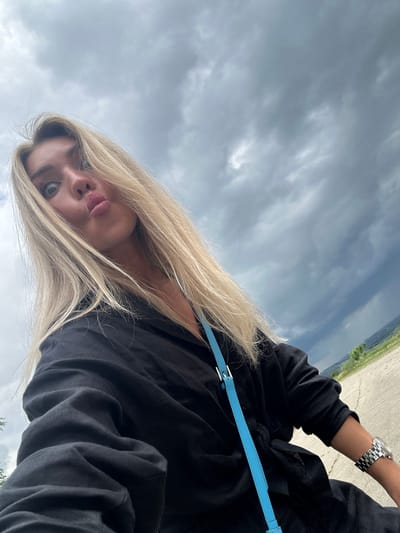A blonde woman in a black shirt with a blue strap bag and a watch on her wrist poses for a selfie outdoors. She has her lips puckered and her eyes wide open. The background is a cloudy sky and a landscape of rolling hills. The location is Krasnaya Polyana, Russia.