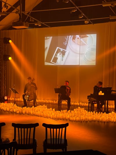 This image depicts a performance venue, likely in Ilyanskiy selskiy Sovet, Belarus, set up for a musical event. The scene is illuminated by warm, orange stage lighting and a multitude of flickering LED candles arranged on the floor in front of a stage. A large screen is projected onto a beige curtain backdrop, displaying an image of a slice of cake and a framed photograph of a couple.
On the stage, three musicians are performing. To the left, a man is seated, playing a cello. In the center, another man is standing, playing a violin. To the right, a man is seated at a black grand piano. All musicians are dressed in dark attire. In the foreground, empty wooden chairs are visible, suggesting an audience area. The overall atmosphere is intimate and visually striking, with the warm candlelight creating a dramatic effect.