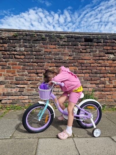 A young girl in a pink hooded sweatshirt, yellow shorts with the word "NOW" printed on them, and pink clogs is leaning over a purple and teal bicycle with training wheels. The bicycle has a purple basket with a purple stuffed toy inside. The girl's face is obscured as she rests her head on the handlebars. The bicycle is parked on a paved walkway in front of a weathered brick wall. Small green plants are growing in the gaps between the bricks and at the base of the wall. The sky above is blue with scattered white clouds. The scene appears to be outdoors, possibly in a residential area of Salford, United Kingdom.