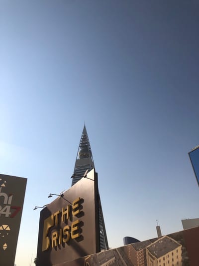 This media file captures an outdoor, urban architectural scene in Riyadh, Saudi Arabia, primarily featuring the distinctive Kingdom Centre Tower and a prominent commercial sign, set against a clear blue sky. The image appears to be taken from a low vantage point, looking upwards, and is presented in an unusual, rotated orientation. If rotated clockwise by 90 degrees for better readability, the sky would be at the top.
The dominant subject is a tall, triangular-shaped skyscraper, the Kingdom Centre Tower, positioned on what would be the right side of the upright frame. Its modern design features glass facades, and glimpses of internal structures or greenery are visible within its upper sections. The tower's pointed apex reaches towards the sky.
In the lower-right area of the upright image, a large, dark-colored sign is prominently displayed. It features the words "THE RISE" in bold, gold, three-dimensional lettering. Small light fixtures are attached along the top edge of this sign, casting shadows that suggest bright daylight. Another partial sign or structure is visible in the lower-left corner, displaying the text "24/7" alongside geometric patterns. Various other building outlines or reflective surfaces can be seen along what would be the left side of the upright image.
The scene is illuminated by bright, clear daylight, indicated by the strong shadows and the expansive, cloudless light blue sky that dominates the upper portion of the image. No people or specific activities are visible, emphasizing the architectural grandeur. The unique shape of the skyscraper serves as a clear location cue, confirming the setting as Riyadh, Saudi Arabia.