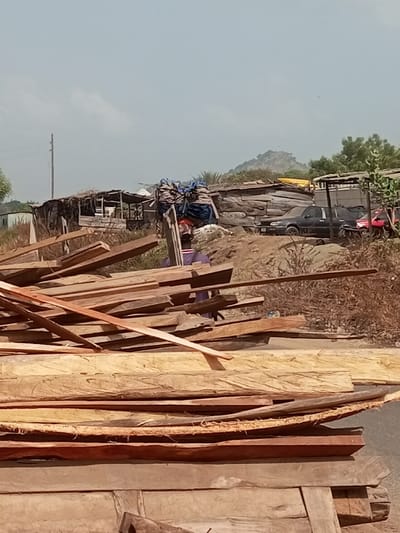 The scene depicts an outdoor environment in Ebiya, Nigeria, under a clear, hazy sky. In the immediate foreground, a substantial, irregularly stacked pile of rough-hewn wooden planks and logs occupies the lower frame, varying in color from light tan to reddish-brown. Behind this lumber, centrally positioned, a person is visible carrying a large, bundled load on their head. The individual wears a dark shirt and a red head covering, with the load appearing to consist of multiple items contained within blue sacks or tarpaulins. The ground beneath them is uneven, reddish-brown earth. In the midground and background, a series of informal structures constructed from various materials, including corrugated metal and wood, are present. Several vehicles are parked among these structures, including a dark-colored sedan and a red vehicle. Sparse, dry vegetation is scattered throughout the area, particularly on the right. A prominent, rocky hill is discernible on the horizon in the distance, against the light blue sky, which also shows a utility pole on the far left.