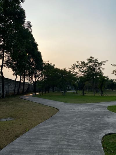 The image depicts a paved pathway winding through a park with lush green grass and trees. A tall, gray concrete wall runs along the left side of the pathway, with trees casting shadows on it. The sky is a soft gradient of pale yellow and blue, suggesting either sunrise or sunset. The pathway forks in the foreground, with one branch curving to the left and the other to the right. This park appears to be located in East Jakarta, Indonesia, a typical urban green space designed for recreation and relaxation. There are no people or animals visible in the scene.