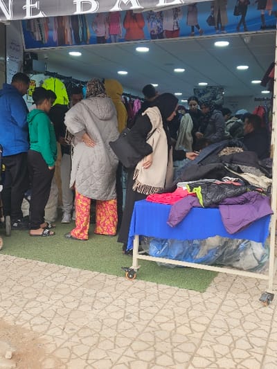 The image portrays an outdoor retail setting in Salé, Morocco, at the entrance of a clothing store. A large banner with white text, partially reading "ONE," and displaying images of children in various outfits, spans the top of the storefront. The store's interior is brightly illuminated by multiple recessed ceiling lights and contains visible clothing racks.

In the midground, multiple individuals are engaged in browsing merchandise. On the left, a person wearing a blue jacket stands with their back to the camera, next to a younger individual in a green hoodie and black pants. To their right, another person in a grey quilted jacket and distinctive pink and yellow patterned trousers also faces away. A woman, dressed in a black abaya and a light beige headscarf, carrying a black shoulder bag, leans over a mobile display table positioned on the right. This table, covered with a blue cloth, holds a pile of assorted garments, including jackets and tops in colors such as pink, purple, black, and lime green. Below the table, several blue plastic-wrapped bundles are visible.

The ground immediately in front of the store entrance is covered by a green artificial turf mat. Beyond this, the ground transitions to light-colored paving stones adorned with an intricate star-like geometric pattern.