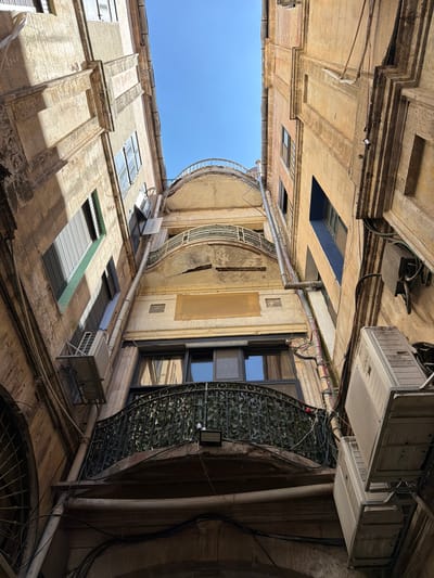 The image captures an upward view of an urban alleyway or courtyard in Jerusalem, Israel, under a clear blue sky. The architecture is characterized by weathered stone buildings with multiple stories, balconies, and numerous windows. Several windows are covered with metal shutters, some of which are partially open, allowing light to stream through.
Prominent objects include multiple air conditioning units mounted on the exteriors of the buildings, some appearing old and somewhat grimy. Various pipes and electrical conduits are visible running along the building facades, indicating the infrastructure of the urban environment. Decorative wrought-iron railings adorn some balconies.
The lighting suggests it is daytime, with strong sunlight casting shadows, highlighting the textures and aged appearance of the stone and plasterwork. The scene conveys a sense of historical depth and everyday urban life, without any people present. The perspective from below emphasizes the towering nature of the buildings and the narrowness of the space between them. No visible text is present in the image.