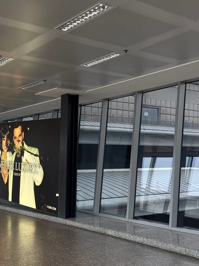 The image captures an interior view of a modern building in Ferno, Italy, looking up towards a ceiling and out through a window, with a large advertisement panel in the foreground.
At the top, a light grey ceiling is visible, featuring a geometric grid pattern formed by recessed panels and beams. Multiple rectangular fluorescent light fixtures, some illuminated, are integrated into the ceiling, along with a small, dark, knob-like object.
Below the ceiling and extending across the middle, a large multi-paned window structure, framed in dark grey or black, offers a view of another building. This exterior building is modern, with a grid-like facade and vertical structural elements. Reflections of the sky and surrounding architecture are visible in its windows. Through one of the windowpanes, a dark door with a small, green sign can be seen on the distant building's facade. A heavy, black horizontal beam runs beneath the lower section of this window structure.
On the right side of the image, a textured, dark grey wall, likely granite, is present, with a distinct lighter-colored trim bordering the window section.
Dominating the lower and central part of the image is a large, brightly lit advertisement. It features a man in a light-colored suit jacket, holding a red rose near his face, gazing towards the viewer. A woman, partially visible below him, also in light attire, holds a red rose. The advertisement's background is dark. Prominent text on the ad reads "WE ARE LUXURY," "MANAGEMENT & PROPERTY GROUP," and "PLEIN.COM."
The scene depicts a clean, contemporary architectural environment, possibly a commercial building or an airport terminal (consistent with the Ferno location). Natural daylight appears to be present, indicated by reflections outside, complementing the artificial interior lighting. No specific activity is occurring.