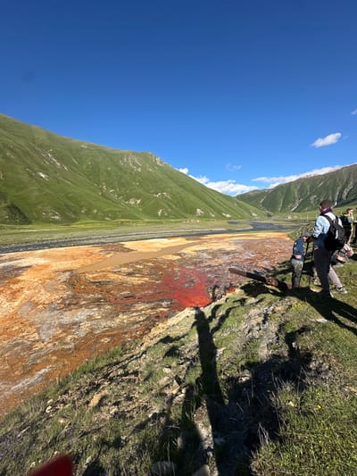 The image depicts a natural landscape with a vibrant, mineral-rich body of water in the foreground. The water exhibits striking patterns of red and orange hues, suggesting the presence of various minerals. Surrounding the water is a landscape of rolling green hills under a clear blue sky, indicative of a mountainous region. Several individuals, appearing to be hikers or tourists, are present on the grassy embankment overlooking the water. They are dressed in outdoor attire and some are equipped with backpacks, suggesting they are engaged in an excursion. One individual is holding a selfie stick. The scene captures a moment of observation and exploration of the unique geological formation. The location is identified as being in Ketrisi, Georgia.
