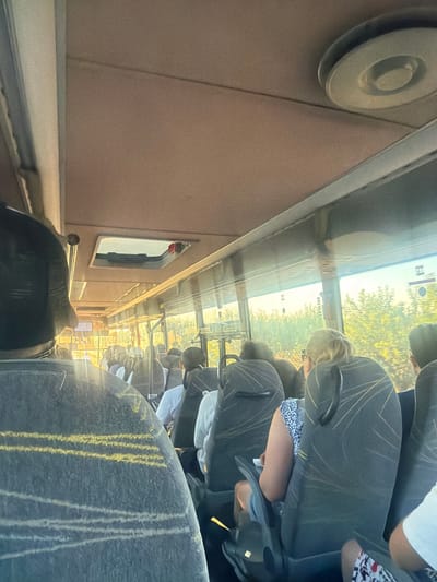 The image depicts the interior of a bus filled with passengers, viewed from a seated perspective. The bus appears to be in motion, as indicated by the sunlight streaking through the windows and the blurred green foliage visible outside. The ceiling of the bus is a light brown color, with a circular vent or light fixture visible in the upper right corner. Several passengers are seated in the frame, mostly facing forward. The seats are dark gray with a pattern of thin gold lines.

In the foreground, a person wearing a dark gray garment with yellow and white striped markings on the back is seated. To their right, a woman with blonde hair tied up is wearing a patterned blue and white top and is holding a white object. Further back, other passengers are visible, some wearing white shirts. The windows offer a view of trees and greenery, suggesting the bus is traveling through a rural or suburban area. A skylight is open in the ceiling, revealing a glimpse of the sky. The overall impression is of a crowded bus journey during daylight.