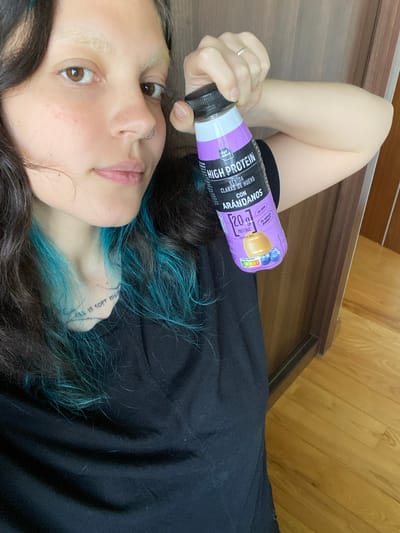 Here's a detailed description of the image:
**Scene:** A selfie taken from a lying down position. The subject is a person with dark hair that is highlighted with teal/blue streaks. They are wearing a black top. They appear to be indoors, on a brown hardwood floor, adjacent to a vertical wooden-paneled wall.
**People:** The primary subject is a person lying down. Their face is partially visible, showing light skin, brown eyes, and a nose ring. A tattoo is visible on their wrist.
**Objects:** The subject is holding a purple bottle labeled "High Protein," with Spanish text including "Arandanos" (blueberries). The bottle contains 20g of protein, and a description of it being made with egg whites and blueberries.
**Actions/Interactions:** The person is holding the protein drink. They are looking at the camera.
**Environment:** The photo appears to be taken inside a room with hardwood floors and wooden walls.
**Location:** It is NOT possible to determine if this image was taken in Villaviciosa, Spain based solely on visual cues. There are no distinguishable landmarks or details in the photo that would pinpoint the location.