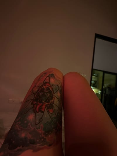 The image shows a person's legs with a tattoo on the left thigh. The tattoo depicts an atom model with celestial elements like stars and nebulae. The person is lying down, with their legs positioned diagonally across the frame. The background is a wall illuminated with a warm, reddish light, and a reflection of a doorway with greenery is visible in a mirror on the right side of the image. The location is identified as Baan Tai, Thailand.