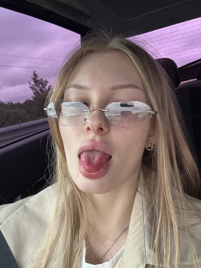 A young woman with blonde hair is pictured inside a vehicle, with her tongue sticking out. She is wearing rimless sunglasses and a light-colored jacket over a white top. A silver necklace is visible around her neck. The background through the car windows shows a blurry landscape of trees and a cloudy, purple sky, suggesting it is daytime. The interior of the car is dark and out of focus, with only the woman and her immediate surroundings clearly visible. The location is identified as Kondratovo, Russia.