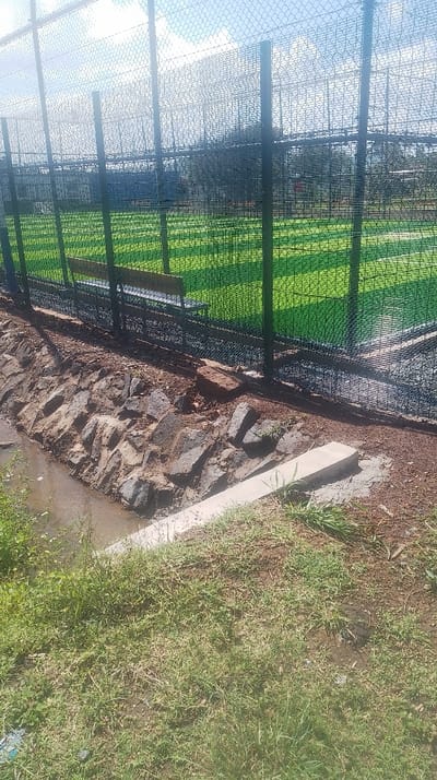 A fenced green space, possibly a sports field, is visible through a wire fence. A small water-filled ditch lined with large stones runs along the base of the fence. A concrete structure is adjacent to the ditch. A grassy area is in the foreground. The day is bright and sunny.