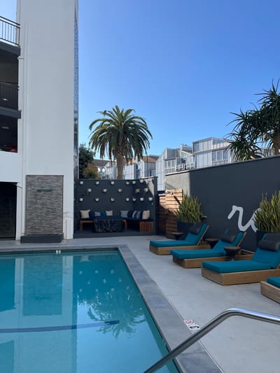 A rooftop pool area displays blue water reflecting a palm tree. Lounge chairs line one side, adjacent to a dark wall adorned with stylized white designs and greenery. An outdoor seating area with dark furniture and white spherical decorations sits beside the pool. Modern architecture is visible in the background under a clear blue sky. The picture is taken from an elevated perspective.