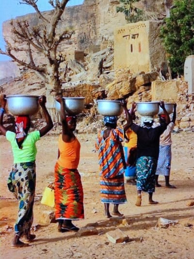 Five women are depicted walking across dry, reddish-brown earth in an arid, rocky area of Dodoma, Tanzania. Each woman carries a large, silver-colored metallic basin balanced on her head.
From left to right:
1. A woman wears a light green short-sleeved top, a patterned long skirt, and a red and white head covering. Her left hand is raised towards the basin.
2. A woman wears an orange sleeveless top and a brightly patterned long skirt in orange, red, green, and black. A yellow bag is visible near her left side. Her left hand is raised towards the basin.
3. A woman, viewed from the back, wears a diagonally striped garment in orange, blue, and white, with a blue patterned head covering. Both hands are raised to the basin.
4. A woman, viewed from the back, wears a black short-sleeved top and a green and white patterned long skirt, with a white head covering. Both hands are raised to the basin.
5. A partially visible woman in the rear wears a light-colored top and a light blue/gray patterned skirt, with a head covering and hands raised to her basin.
The ground is dusty with scattered small rocks. In the background, a prominent leafless tree is visible on the left. Central to the mid-ground stands a light-colored, rectangular building with geometric markings, appearing to be constructed from earth or mud. This structure is surrounded by natural rock formations and additional, less distinct, stone and earth buildings. Sparse green vegetation is present on the far right. The sky is clear and light blue.