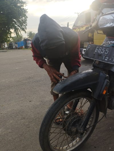 An outdoor, eye-level shot captures a person attending to a motorcycle on a paved surface, likely a roadside in Kecamatan Jangkar, Indonesia. The central figure, possibly male, is clad in a dark hooded top with red sleeves, bending forward with their face obscured by the hood. Their hands, appearing weathered, are engaged with an object, possibly a tool, suggesting they are performing maintenance or repairs. They wear sandals.
In the foreground to the right, the front wheel and fender of a black and silver motorcycle dominate the frame. The license plate is visible, displaying "P 6855" and partially obscured characters "12 7". The motorcycle shows signs of wear, with some rust on the spokes and hub.
The background reveals a mix of natural and man-made elements. To the left, large green trees are visible, along with several informal stalls or structures covered with blue tarpaulins, indicative of a market or roadside vendor area. Behind the person and the motorcycle, a large yellow and red truck is partially visible, bathed in bright sunlight. The low angle of the sun suggests either early morning or late afternoon, creating a bright glare on the right side of the image and illuminating the scene with a soft, warm light. The sky above is partly cloudy. The overall impression is one of everyday activity in a roadside or semi-urban setting.