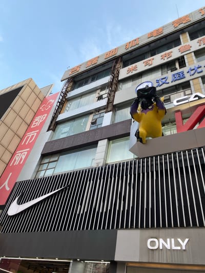 This is a low-angle, eye-level shot of a building facade in Jinjiang District, China. The building has multiple signs, including a large red banner with white Chinese characters and a butterfly logo, and the Nike logo on a black and white striped facade. Atop the building, a bright yellow inflatable figure in a yellow hazmat suit and a black helmet is visible. The building has several floors with windows and some visible signage in Chinese characters. The sky is blue and clear.
