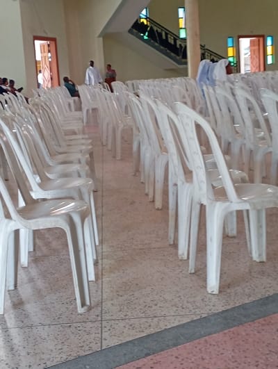 The scene is located at the coordinates -3.373, 36.694, which corresponds to a location within the city of Arusha, Tanzania. The image is taken inside a large, open space with a tiled floor. The space is filled with rows of white plastic chairs, arranged in straight lines. In the background, there is a stairway leading to an upper level, a partially obscured door on the left side, and another door on the right side with a stained glass window. There are four people in the scene, three standing in the background and one sitting in a chair in the middle of the image. One of the people in the background is wearing white clothing.  The image is likely taken during a gathering or an event, as the chairs are set up in rows, suggesting that it is a place for people to sit and watch something.