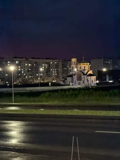 An evening view of a residential area and a church in Nizhnekamsk, Russia. The scene is illuminated by streetlights and the lights of apartment buildings. A multi-story apartment building with many illuminated windows dominates the left side of the frame. To the right of the apartment building is a white church with a golden dome and bell tower, also illuminated. A grassy median separates the foreground road from the buildings. The road in the foreground is wet and reflects the ambient light. Two vertical silver poles are visible on the right side of the road. The sky is dark and appears to be twilight.