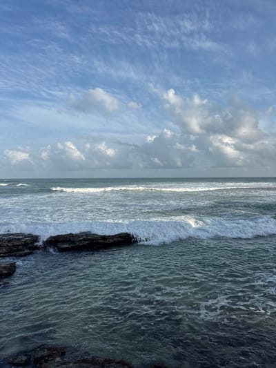 The image depicts a view of the ocean from the shore in Goviyapana, Sri Lanka. Dark, wet rocks are visible in the foreground, partially submerged by the incoming tide. Waves are breaking over these rocks, creating white foam. The water is a deep blue-green, with ripples and foam indicative of movement. In the mid-ground, several lines of waves are rolling towards the shore, some with crests of white foam. The horizon is visible, separating the ocean from the sky. The sky is a pale blue with scattered, wispy white clouds, some of which are tinged with grey, suggesting a partly cloudy day. The overall scene is one of a dynamic coastal environment under a bright sky. There are no people or animals present in the image.