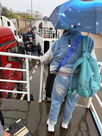 The image captures a rainy scene on what appears to be a vehicle and passenger ferry. In the foreground, a person is seen from behind, dressed in a denim jacket over a grey hoodie, blue jeans rolled at the cuffs, and white sneakers. They wear a teal-colored backpack and a bright blue strap with a buckle across their chest. The person is holding a dark blue umbrella with a galaxy or cosmic print, featuring the word "PLANET" written vertically.

The ferry deck is wet, indicating rain or recent precipitation, with visible ripples on the surface of the water near the edge. White metal railings separate the foreground from the midground. Below the railings, dark water is visible.

In the midground and background, numerous other people, many carrying bags and some holding umbrellas, are gathered on the ferry deck, likely waiting or boarding/disembarking. Several large trucks are also on board, including a prominent red truck with "J6P" and Chinese characters ("解放 FAW" and other smaller text) visible on its side, and a white truck. In the distant background, a blue bus is visible, and there are buildings along a dock or pier, suggesting a port or ferry terminal. The overall atmosphere is overcast and damp.