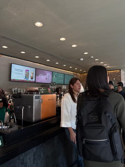 The scene depicts the interior of a Starbucks coffee shop located in San Agustín, Mexico. In the immediate foreground, the back of a person wearing an olive-green long-sleeved garment and a black backpack with a metallic "AB" monogram is visible. To the left of this individual, a woman with dark brown hair, wearing a white long-sleeved shirt and blue jeans, stands facing right, looking off-camera. Her left hand, adorned with rings, is partially visible.

Behind these individuals, a long, dark speckled countertop runs horizontally, forming the service area. On the counter, a large stainless steel and copper-toned espresso machine is prominent, with a clear glass bowl filled with roasted coffee beans situated on its top. To the far left, a barista in a green apron is partially visible behind the counter, near a stainless steel sink and faucet.

Above the service counter, three digital display screens are mounted on a dark grey wall. The screens show various promotions and menu items in Spanish, including "Tostado ligero y notas a miel" and "NUEVO Chai Cherry." A section of the wall to the right features a decorative, stylized brown and tan pattern. In the background, several blurred figures of other patrons or staff are discernible. The ceiling is a textured, light grey concrete, equipped with numerous recessed circular spotlights providing illumination, along with a white smoke detector and a small security camera. The environment indicates a contemporary and functional coffee shop setting.