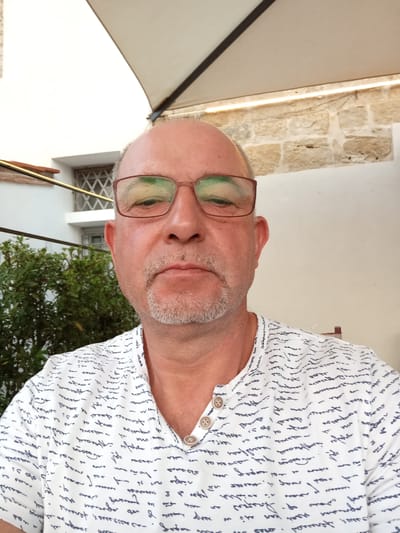 A selfie of a man with a bald head, wearing glasses and a white shirt with a dark handwritten script pattern. He has a goatee and is looking directly at the camera. The background shows a white awning, stone walls, and green foliage. The scene appears to be outdoors in an urban setting, possibly a patio or courtyard. The man is the sole subject.