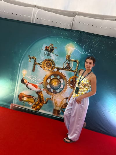 In Málaga, Spain, a young woman stands on a red carpet in front of a large, detailed mural. The mural depicts a steampunk-inspired scene within a glass dome. Gears, pipes, and lightbulbs are prominent elements, with figures engaged in various activities. One figure appears to be falling with a lightbulb on his head, while another is riding a contraption resembling a bicycle with wings. The woman is wearing a yellow camisole with a lemon print and light purple satin pants, along with black flip-flops. She has a lanyard with badges around her neck. The background of the mural is a dark teal with white speckles resembling stars and stylized lightning. The environment suggests an indoor event or exhibition, with professional lighting illuminating the scene.
