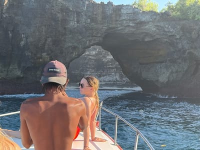 In Nusa Penida, Indonesia, two individuals are on a boat, with a dramatic natural rock arch formation in the background. The person in the foreground, viewed from behind, wears a baseball cap and appears to be a shirtless male. To their right, a woman with blonde hair tied back, wearing sunglasses and a swimsuit, leans forward on the boat's railing. The boat is navigating through choppy, dark blue water, with the sun casting warm light on the people and the rocky cliffs. The rock arch, a prominent feature of the landscape, reveals a sandy beach or shallow water within its opening. Vegetation is visible growing on the cliff tops.