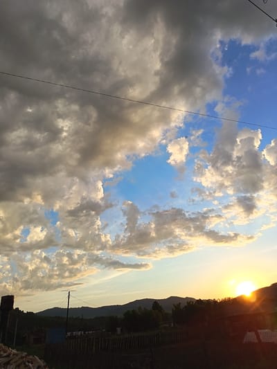 The image depicts a rural landscape at sunset in Soloneshnoye, Russia. The sky is filled with dramatic clouds, ranging from dark gray to bright white and tinged with golden hues from the setting sun. A power line stretches horizontally across the upper portion of the frame. The foreground shows a dark, silhouetted rural village with trees and a wooden fence. In the far right of the frame, the sun is setting behind a hill, casting a bright glare and orange light. To the left, a water tower and a pile of firewood are visible. The overall impression is one of a peaceful, yet dynamic, rural evening.
