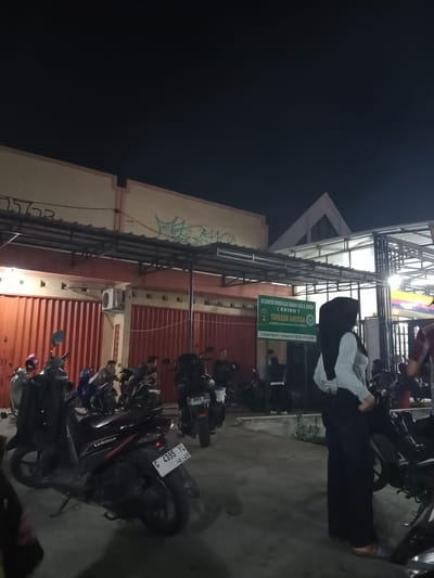 This media file depicts an outdoor scene at night, likely in Sekopek Wetan, Indonesia, as indicated by the visible address on a green sign. The setting appears to be a parking area or an open space in front of a building, with several motorcycles parked closely together. The building in the background has a corrugated metal facade in red and a lighter colored upper section with some graffiti. Above this structure, a modern-looking roofline is visible against the dark night sky.
The primary subjects are several people, some of whom are standing near or sitting on motorcycles. One individual in the foreground is wearing a black hijab and a white top, with dark pants. Their posture suggests they might be conversing or observing something. Other individuals are less clearly defined due to the low light conditions, but they appear to be casually gathered around the parked vehicles.
A prominent green sign displays text in Indonesian, identifying "KELOMPOK BIMBINGAN IBADAH HAJI & UMROH (KBIHU) YAYASAN AROFAH". Below this, an address and phone number are visible. The time of day is clearly night, suggested by the darkness of the sky and the artificial lighting illuminating the scene. No specific weather conditions are evident.
Several motorcycles are parked, including a red and black scooter in the foreground with a license plate reading "G 4395 YL 10.29". Another motorcycle in the background has a license plate "H 2491 BVD 61-30". The overall atmosphere appears to be relaxed and casual, with people gathering in the evening.