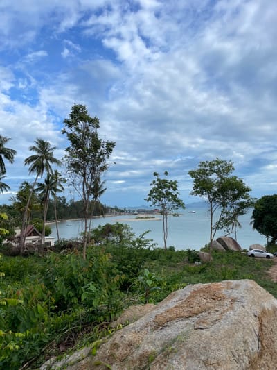 From an elevated perspective on a grassy slope, a partial view of Baan Wok Tum, Thailand, unfolds on a partly cloudy day. A substantial rock dominates the foreground's right, its textured surface in clear view. Lush greenery and trees fill the slope leading down to the water's edge. Palm trees and other trees are interspersed along the slope. A white car is parked near the top of the slope, in front of a second large rock. The water is visible in the distance, with land faintly outlining the horizon. The sky is mostly obscured by clouds.