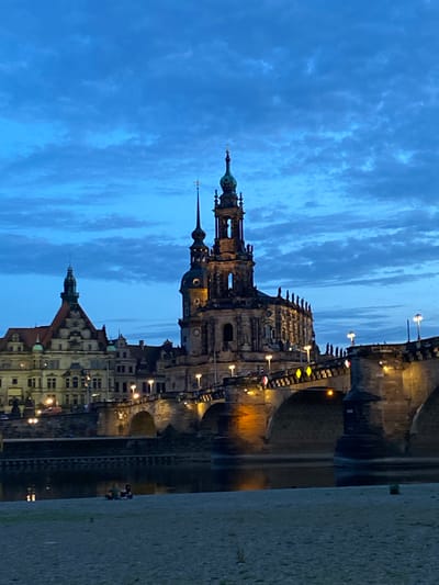 The image shows the city of Dresden, Germany at dusk. The Frauenkirche church is prominently featured, with its distinctive dome and spires. The Elbe River runs through the city, reflecting the lights of the buildings and bridge. The Augustus Bridge is visible, connecting the old town with the new town. The sky is a deep blue with scattered clouds. The buildings are illuminated with warm lights, highlighting their architectural details. Some pedestrians are visible on the riverbank.