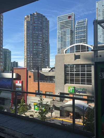 This is a medium shot, low-angle, vertical photograph of a city skyline on a clear, sunny day. The scene is dominated by several modern skyscrapers, characterized by their glass facades and intricate architectural designs. In the mid-ground, a Holiday Inn Express hotel is visible, its green and white logo prominently displayed. Other commercial establishments, including a Subway restaurant, a Best Buy, and a Rexall pharmacy, are also present, suggesting a bustling urban environment. The image's perspective looks up at the buildings, emphasizing their height and grandeur. The sky is a vibrant blue, with a few wispy clouds adding texture. The presence of a sidewalk and street suggests a public area. The overall impression is one of a vibrant and modern city center, possibly during the daytime given the bright sunlight. The visible text includes brand names such as "Holiday Inn Express," "SUBWAY," "BEST BUY," and "Rexall," as well as street addresses, indicating a commercial district. There are no people clearly visible in the frame, but a single person can be seen walking in the distance near the Holiday Inn Express. The metallic structures in the foreground suggest the photograph was taken from an elevated viewpoint, possibly a balcony or walkway. The composition highlights the interplay of architectural styles and the urban landscape.