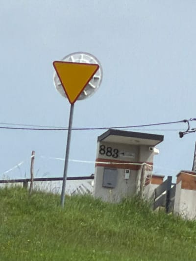 The image depicts an outdoor scene with a yield sign prominently displayed on a metal pole. The sign is triangular, with a yellow interior and a red border. Behind the sign, a portion of a small, light-colored building is visible. This building has a dark, angled roof and features the numbers "883" in black on its side. A small, gray rectangular object, possibly a vent or a small window, is also visible on the building's facade, along with a white box-like fixture. To the right of this building, a dark, angled fence or gate structure is present. The scene is set on a grassy hillside, with a few thin, dry branches or small trees also visible in the background. The sky is a pale blue with a slight haze. The overall impression is that of a rural or semi-rural location, possibly on the outskirts of a town or village. Based on the provided information, this scene is located in Męcina, Poland.