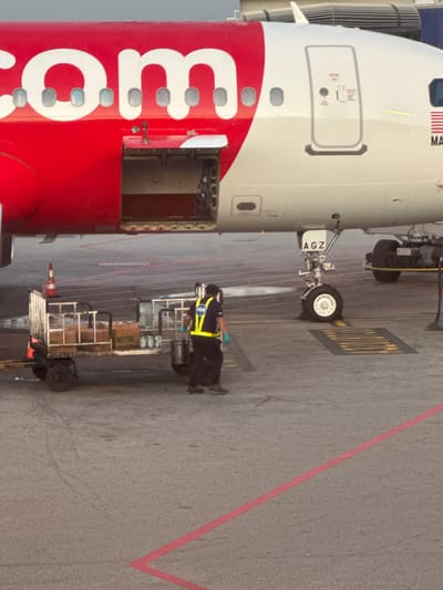 An exterior, eye-level shot captures a portion of a red and white passenger airplane at the Sepang, Malaysia airport. The word "com" is emblazoned in large white lettering on the red portion of the aircraft's fuselage. A cargo door is open, revealing the dark interior. Below the aircraft, a baggage cart is loaded with boxes and what appear to be canisters. A worker wearing a high-visibility yellow vest with "ADE" printed on it, dark pants, and gloves is standing next to the cart. They are holding a black bucket and appear to be in the process of unloading it. The ground is asphalt, marked with red lines. The plane's landing gear is visible, as are other airport structures in the background.