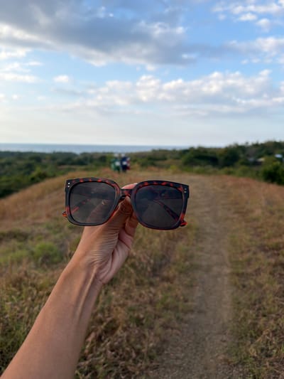 In the foreground, a person's left forearm and hand are visible, holding a pair of tortoiseshell-patterned sunglasses. The sunglasses are positioned slightly to the right of the frame, with their lenses facing forward. The background depicts a natural landscape, characterized by dry, grassy hills and a dirt path leading into the distance. In the far background, a body of water is visible, likely the ocean, meeting a hazy horizon under a sky filled with white and grey clouds. Further into the landscape, two individuals can be seen walking on the path in the distance. The scene suggests an outdoor setting, possibly a hiking trail or a vantage point overlooking the coast. The overall impression is one of a serene, natural environment.