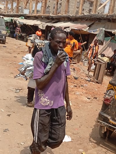 A man drinks from a pouch in a crowded outdoor market. He wears a purple shirt with a white floral design, black shorts with white stripes, and sandals. A gray cloth is slung over his shoulder. Behind him are market stalls with people, a black van, and a person carrying a large bundle. To his right, part of a vehicle with yellow stripes is visible. The ground is dirt with scattered debris. The background shows a building under construction or partially demolished.