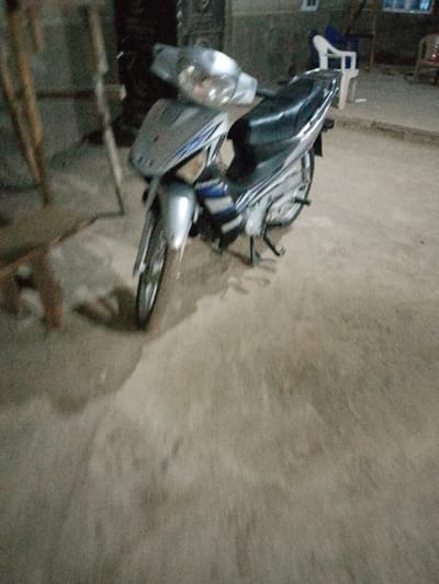An overhead medium shot shows a silver and black motorcycle parked on a dirt ground. The motorcycle is facing left, with its front tire closest to the viewer. The motorcycle has a silver front fairing with blue and white accents. The seat is black. The background is blurred and appears to be an outdoor area, possibly a street or yard, with some wooden structures and furniture visible. The overall lighting is dim, suggesting it might be evening or nighttime. The location is identified as Serti, Nigeria.