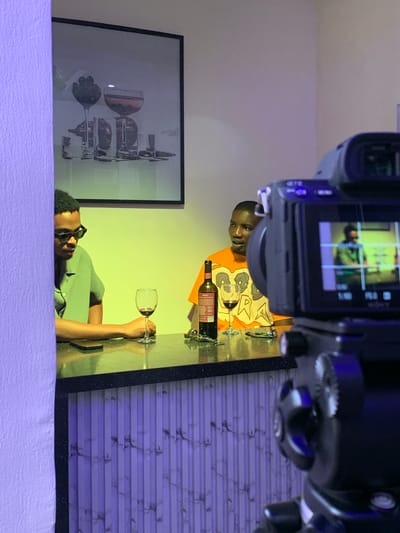 An indoor scene in Moba, Nigeria, depicts two adult males seated at a dark, reflective countertop, being filmed by a foreground camera. The man on the left, wearing black rectangular sunglasses and a light green collared shirt, sits with his left arm resting on the counter. A black smartphone and a wine glass containing red liquid are positioned in front of him. He is oriented towards the right. The man on the right, wearing an orange t-shirt with a graphic design, has a dark wine bottle with a red and white label, and another wine glass containing red liquid, in front of him. He is oriented towards the left, appearing engaged in conversation. The background wall is illuminated with a yellow-green light, featuring a large framed picture depicting stylized glassware. The immediate foreground wall to the left and the base of the counter exhibit vertical ribbed panels with a marble pattern, illuminated by a purple light. A black video camera, positioned in the lower right foreground, points towards the subjects, with its flip-out screen displaying a live feed of the man in the green shirt.