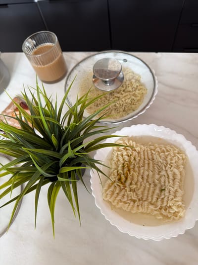 The image shows a close-up view of two bowls of instant noodles and a glass of tea on a white marble countertop. One bowl of noodles is covered with a clear glass lid with a silver handle. The other bowl of noodles is uncovered and partially submerged in broth. A small artificial green plant is positioned to the left of the bowls. In the background, there are black kitchen cabinets and a sliver of a silver appliance. The lighting is bright and appears to be from an overhead source. The scene is set in what appears to be a kitchen environment. There are no people or animals visible in the image. The location is described as being in the city of Семилуки, Russia, though this context cannot be visually verified from the image itself.