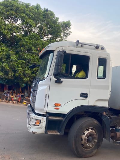A white truck is parked on a road in Abofifia, Nigeria. The driver, wearing a yellow shirt, is visible inside the cab. A tree stands next to the truck. People and businesses line the street.